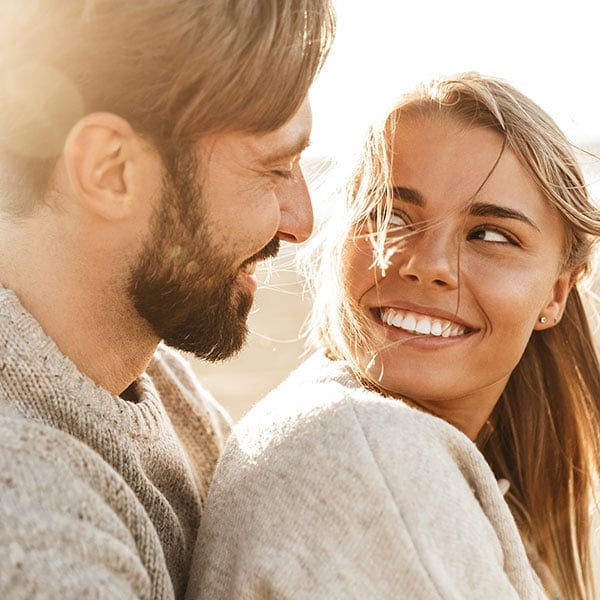 A couple smiles at each other outdoors, sharing an affectionate moment in warm sunlight.